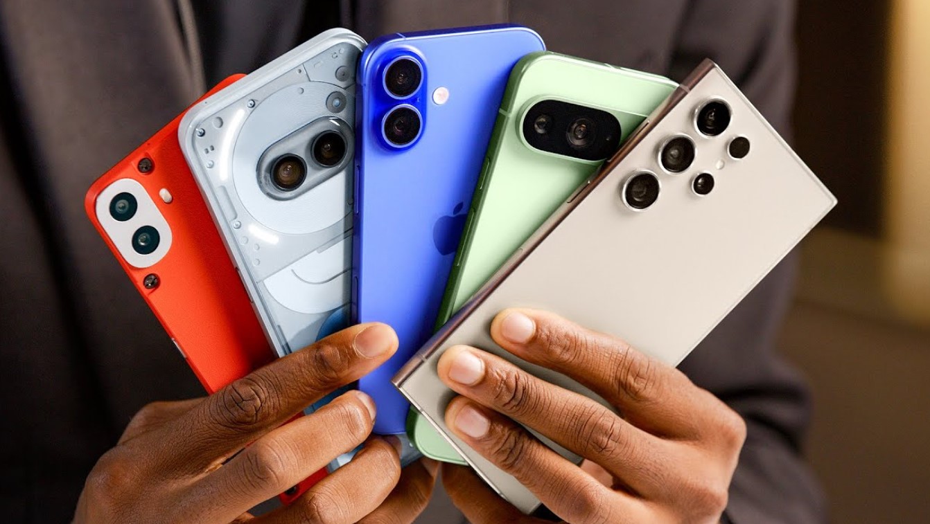 من iPhoneIslam.com، شخص يحمل خمسة هواتف ذكية مختلفة، كل منها بلون فريد: الأحمر والأبيض والأزرق والأخضر والبيج، ومن بينها هاتف آي فون 16 بتصميم كاميرا مميز يميزه عن الباقي.