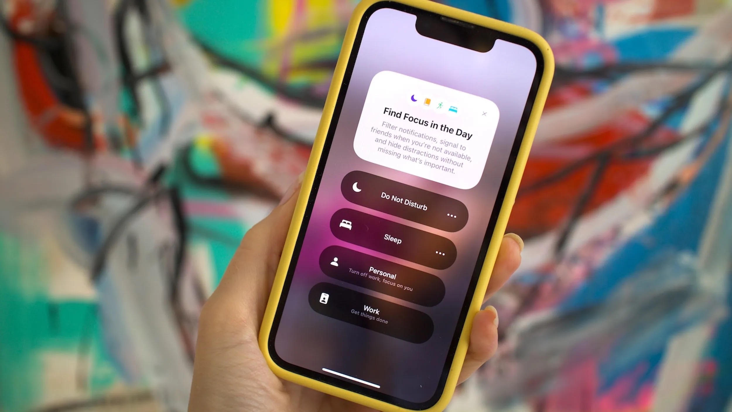 من iPhoneIslam.com، تحمل يد هاتف آيفون يعرض قائمة وضع التركيز مع خيارات مثل عدم الإزعاج والنوم والشخصي والعمل. الخلفية ملونة ومجردة، مما يعزز التصميم الأنيق لهذا الجهاز الاحترافي.