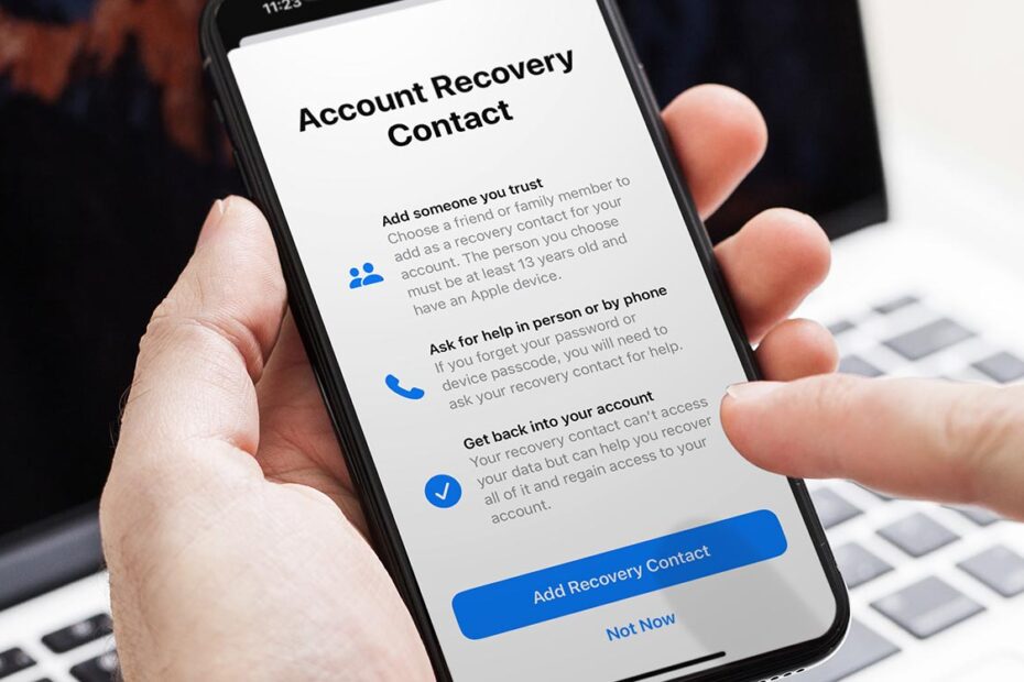 من iPhoneIslam.com، يد تحمل هاتفًا ذكيًا تعرض شاشة "جهة اتصال استرداد الحساب"، وتعرض خيارات مثل إضافة جهة اتصال مثل الحساب للمساعدة في استعادة حساب Apple واستعادة الوصول إلى الحساب.