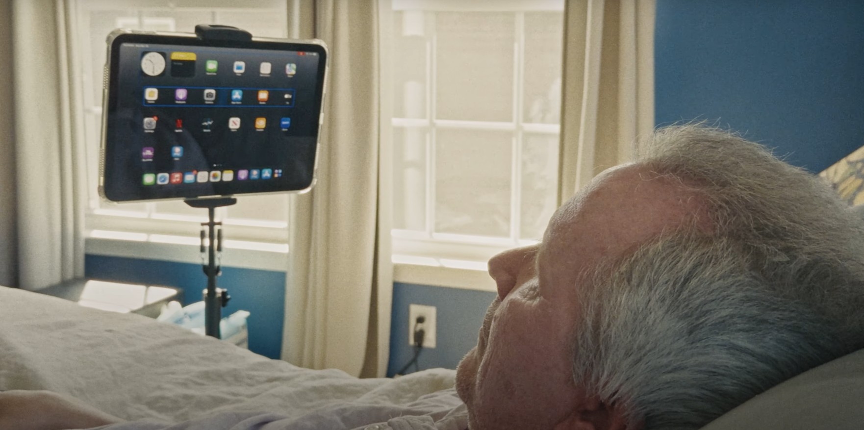 من iPhoneIslam.com، رجل مسن مستلقٍ على السرير، ينظر إلى جهاز لوحي مثبت على حامل، ويعرض شاشة رئيسية مع أيقونات تطبيقات مختلفة في غرفة مضاءة بنور الشمس.
