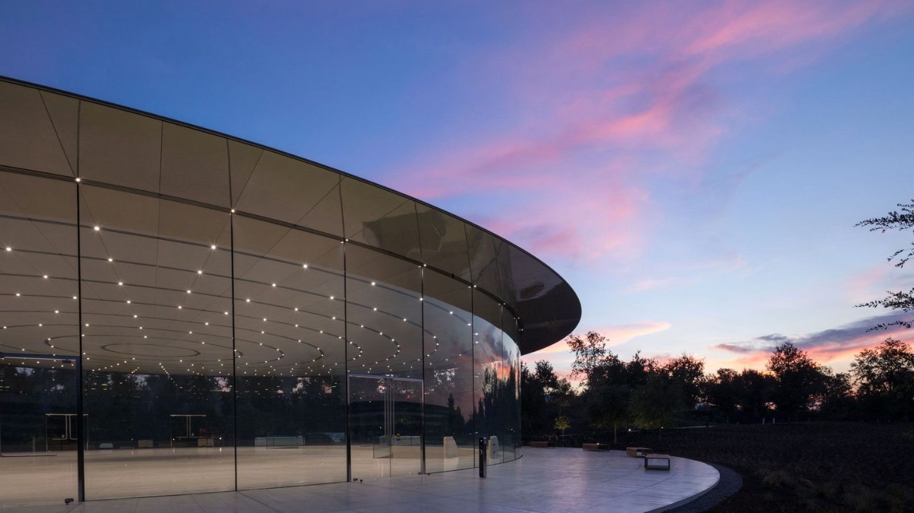 من iPhoneIslam.com، يظهر مبنى زجاجي حديث ذو سقف منحنٍ وسقف مضاء عند الغسق، مع أشجار وسماء ملونة في الخلفية - مما يهيئ المسرح لحادث آيفون 17.