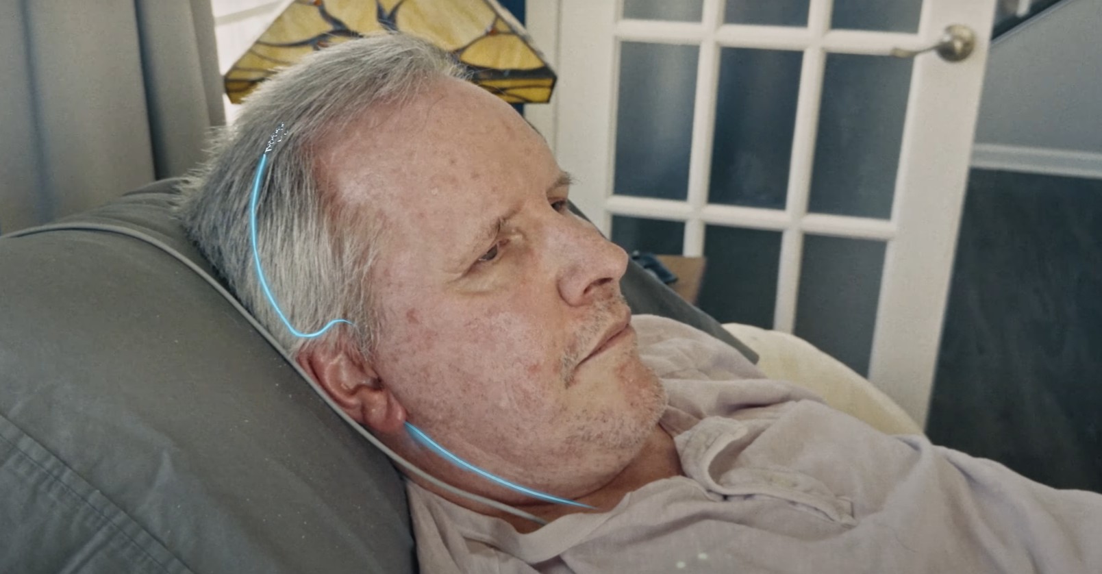 من iPhoneIslam.com، رجل مسن ذو شعر خفيف متكئ على وسادة ويرتدي قميصًا شاحبًا وسماعات رأس سلكية وهو يستمتع بالإدارة في غرفة ذات باب زجاجي ومصباح زجاجي ملون في الخلفية.