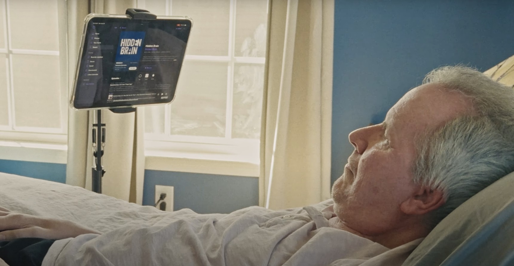 من iPhoneIslam.com، رجل مسن يستلقي في السرير ويشاهد الآي-آي-آي-آي مثبت على حامل، مع ضوء الشمس القادم من خلال نافذة خلفه.