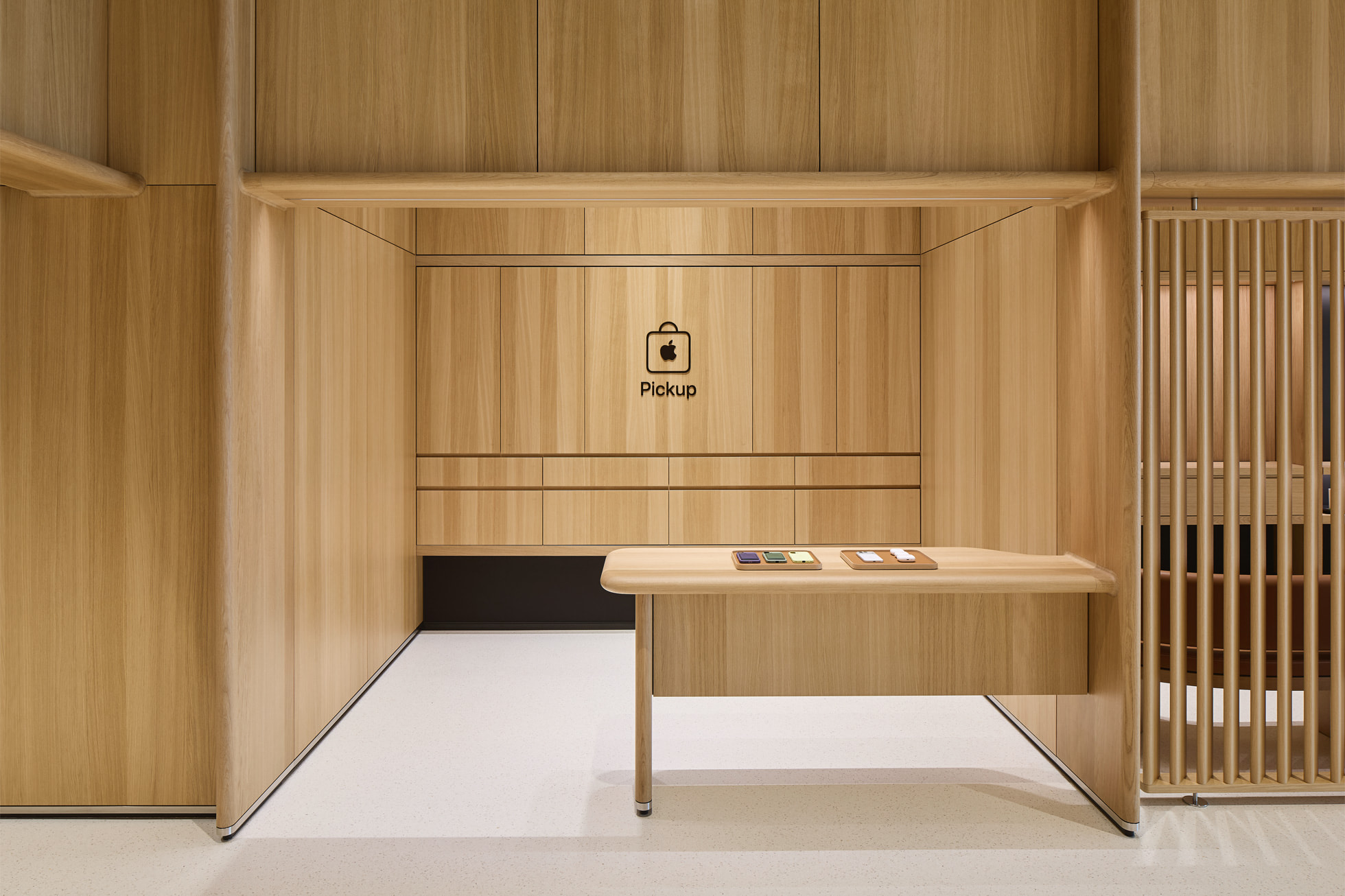 من موقع فون إسلام: منضدة خشبية بسيطة في متجر آبل مع شعار Apple على الحائط وطاولة خشبية بسيطة تعرض ثلاث قطع تقنية صغيرة, تعكس أسلوب العلامة التجارية المميز في الإمارات العربية المتحدة.