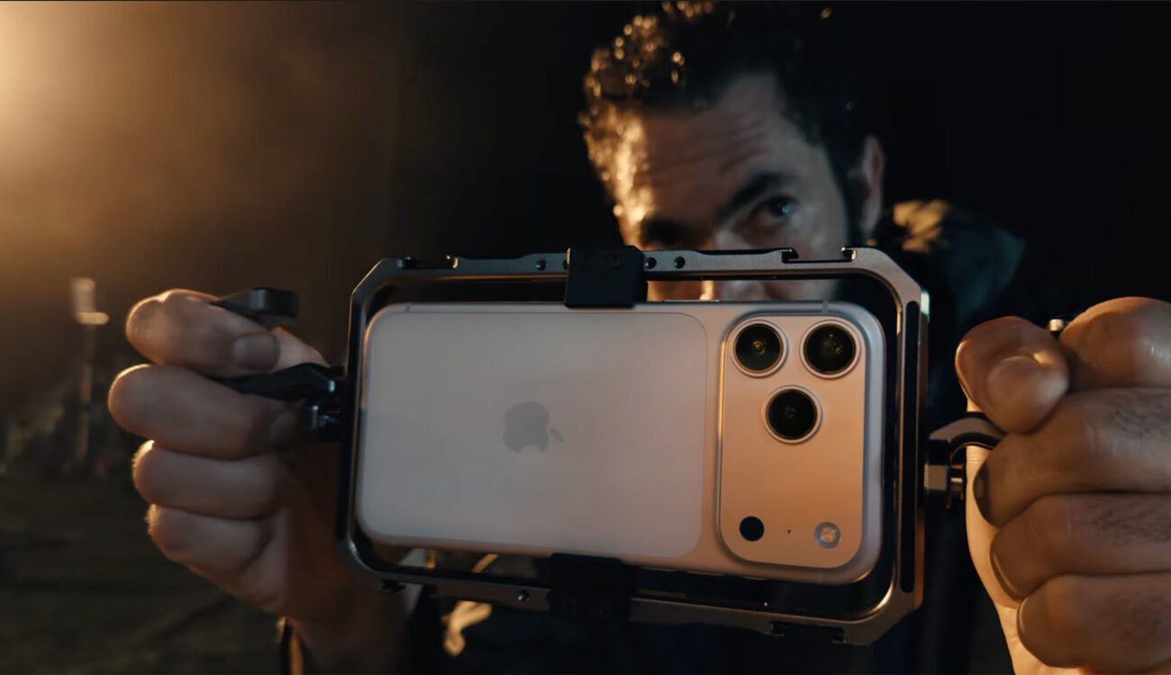 من موقع فون إسلام: شخص يمسك بجهاز أبيض آي-فون 17 برو كاميرات آيفون ثلاثية مثبتة في جهاز معدني، يستعد للتصوير أو التقاط صورة في بيئة خافتة الإضاءة.