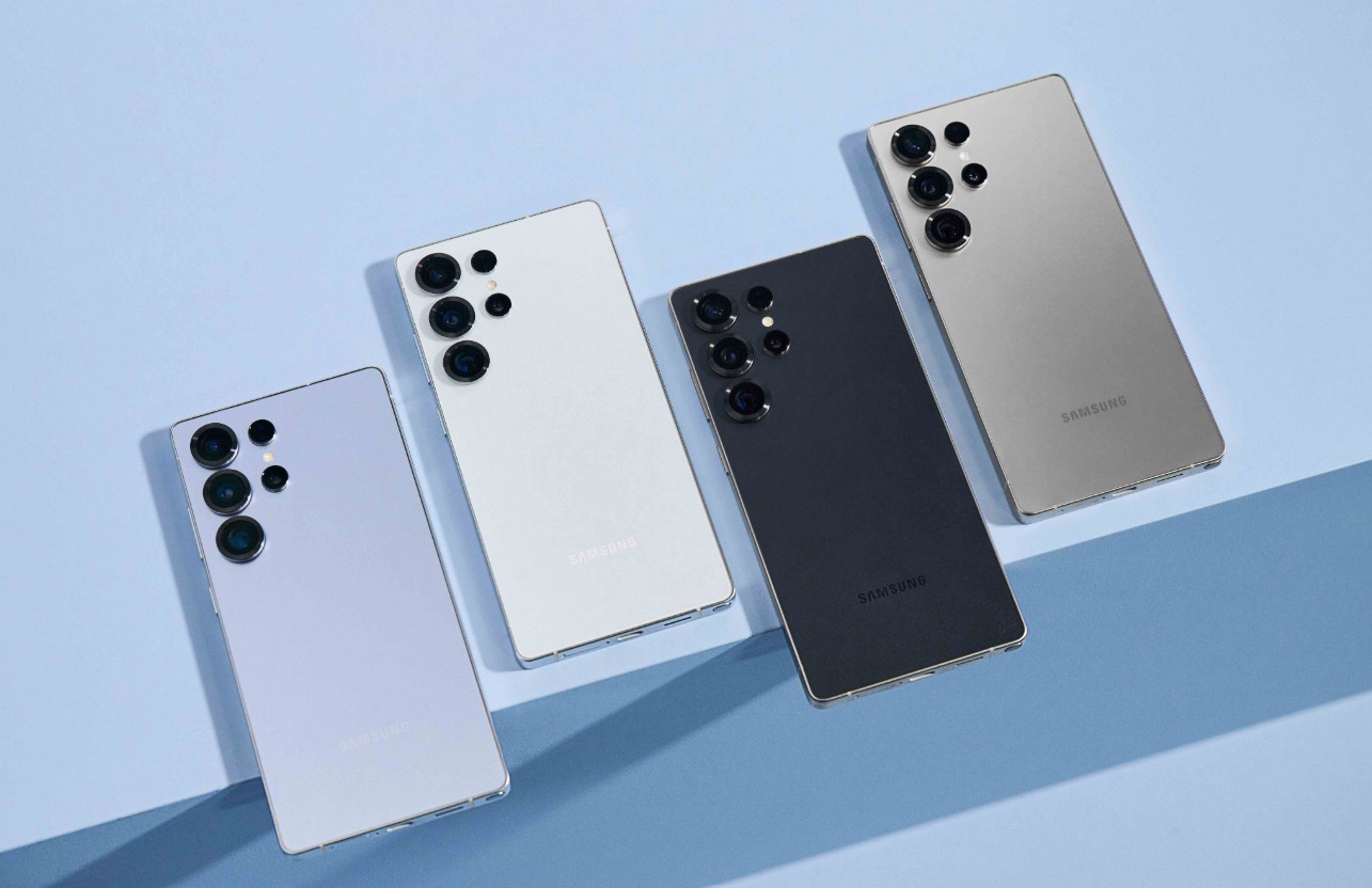 من موقع فون إسلام: أربعة هواتف ذكية، بما في ذلك جالاكسي إس إس 25 ألترا باللون الأبيض والأرجواني الفاتح والأسود والذهبي، مرتبة بشكل مائل على سطح أزرق فاتح.