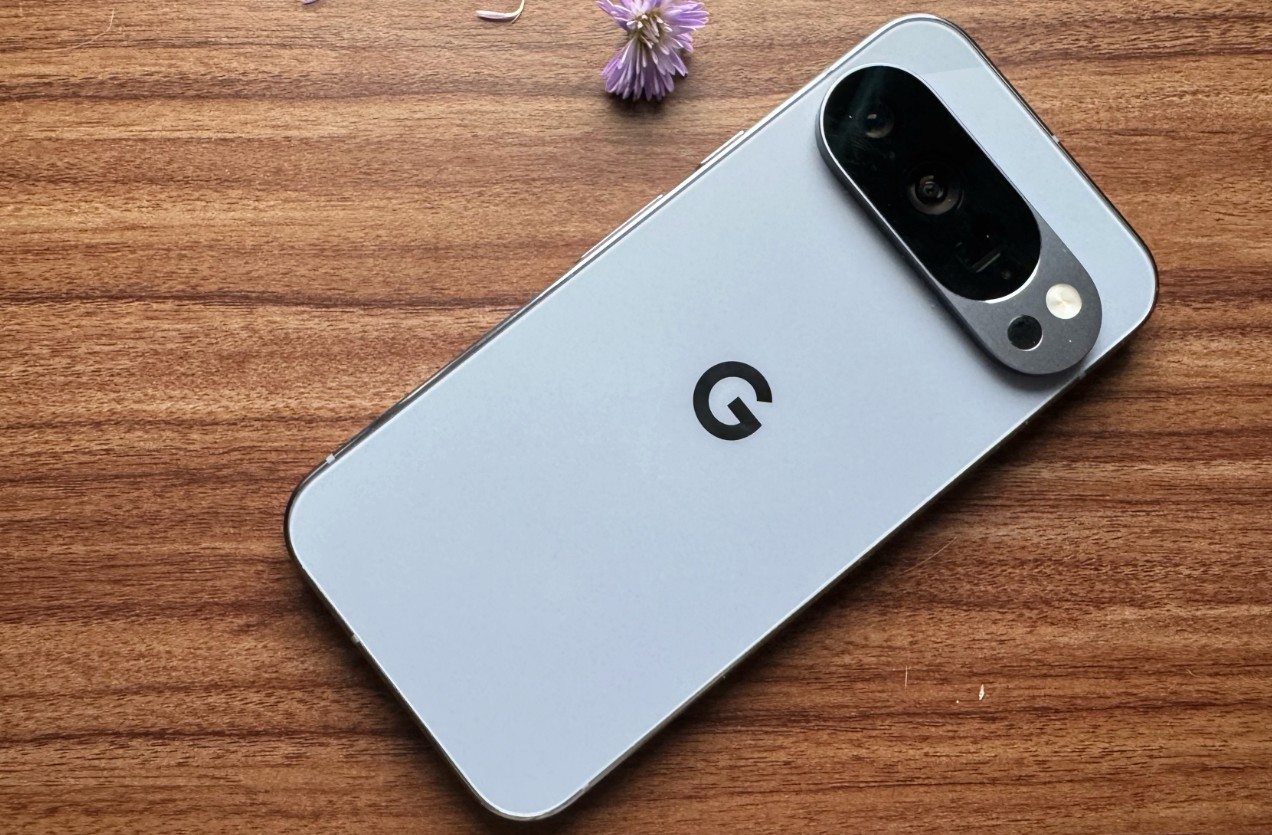 من موقع فون إسلام: هاتف ذكي أبيض اللون بيكسل 10 برو هاتف ذكي مزود بوحدة كاميرا خلفية كبيرة ملقى على سطح خشبي بجانب زهرة أرجوانية صغيرة.