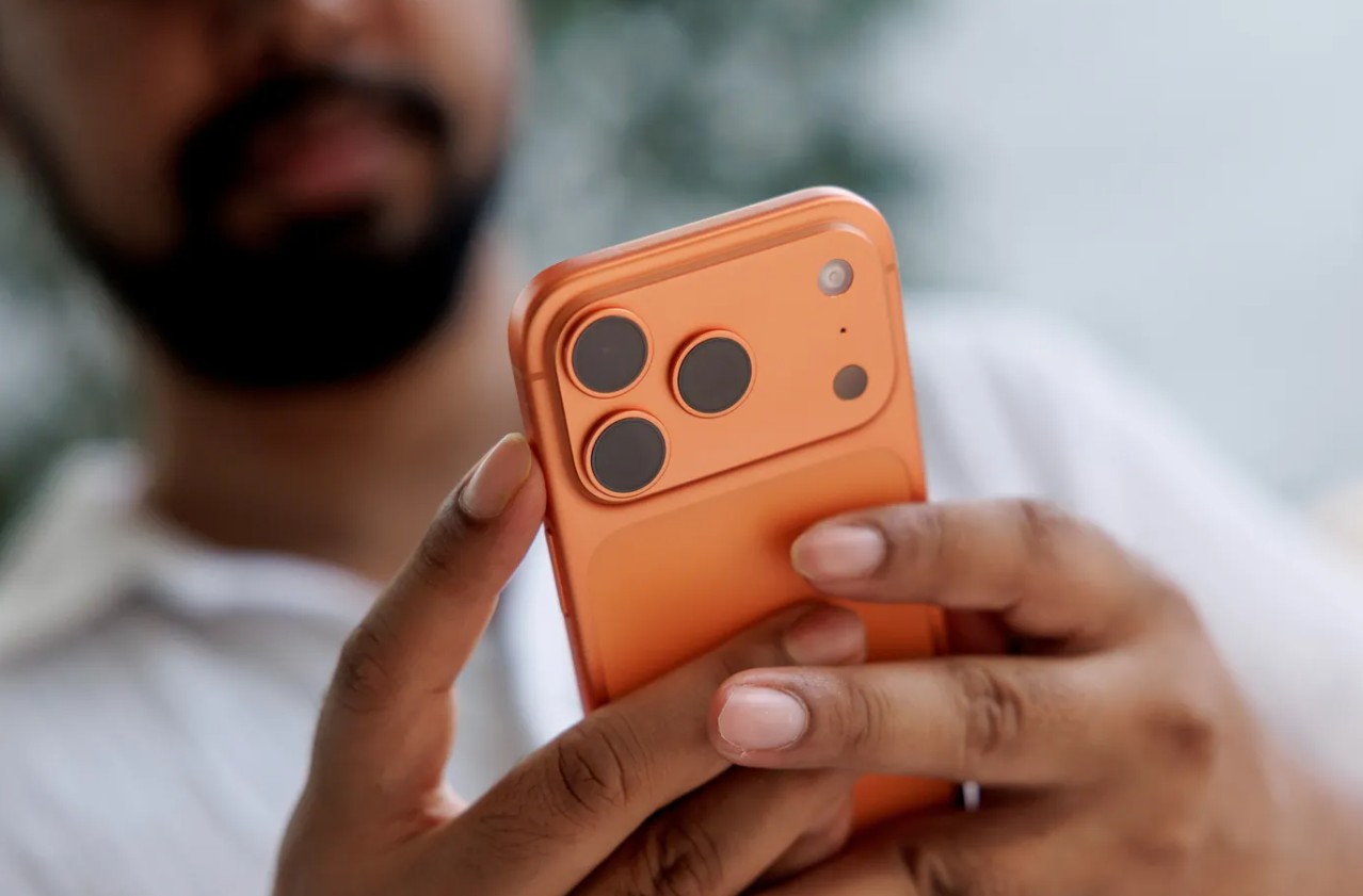 Da Phone Islam: una persona che tiene in mano e utilizza uno smartphone arancione con tre fotocamere posteriori e una custodia arancione abbinata attiva Siri per avviare una conversazione ChatGPT.