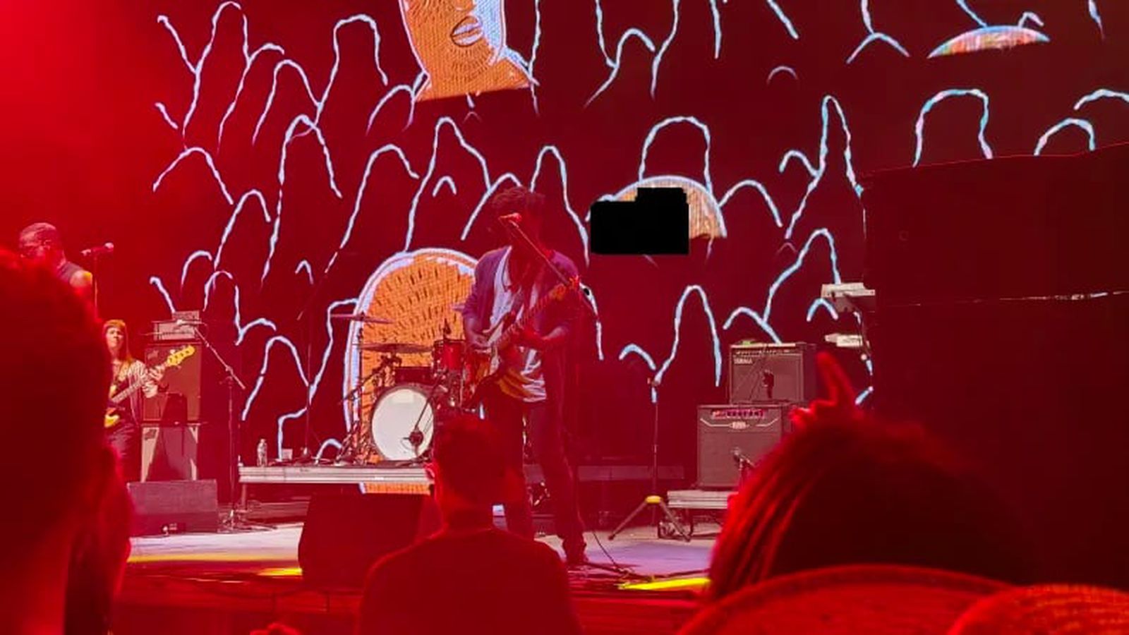 من موقع فون إسلام: تؤدي فرقة موسيقية عرضاً على المسرح تحت إضاءة حمراء مع عرض فن الخطوط البيضاء المجردة والأشكال البرتقالية على شاشة في الخلفية، مما يخلق مشهداً نابضاً بالحياة ومثالياً لمشاركة تحديثات التكنولوجيا الجديدة أو تحديثات ملخصات ملخصات أسبوعي.