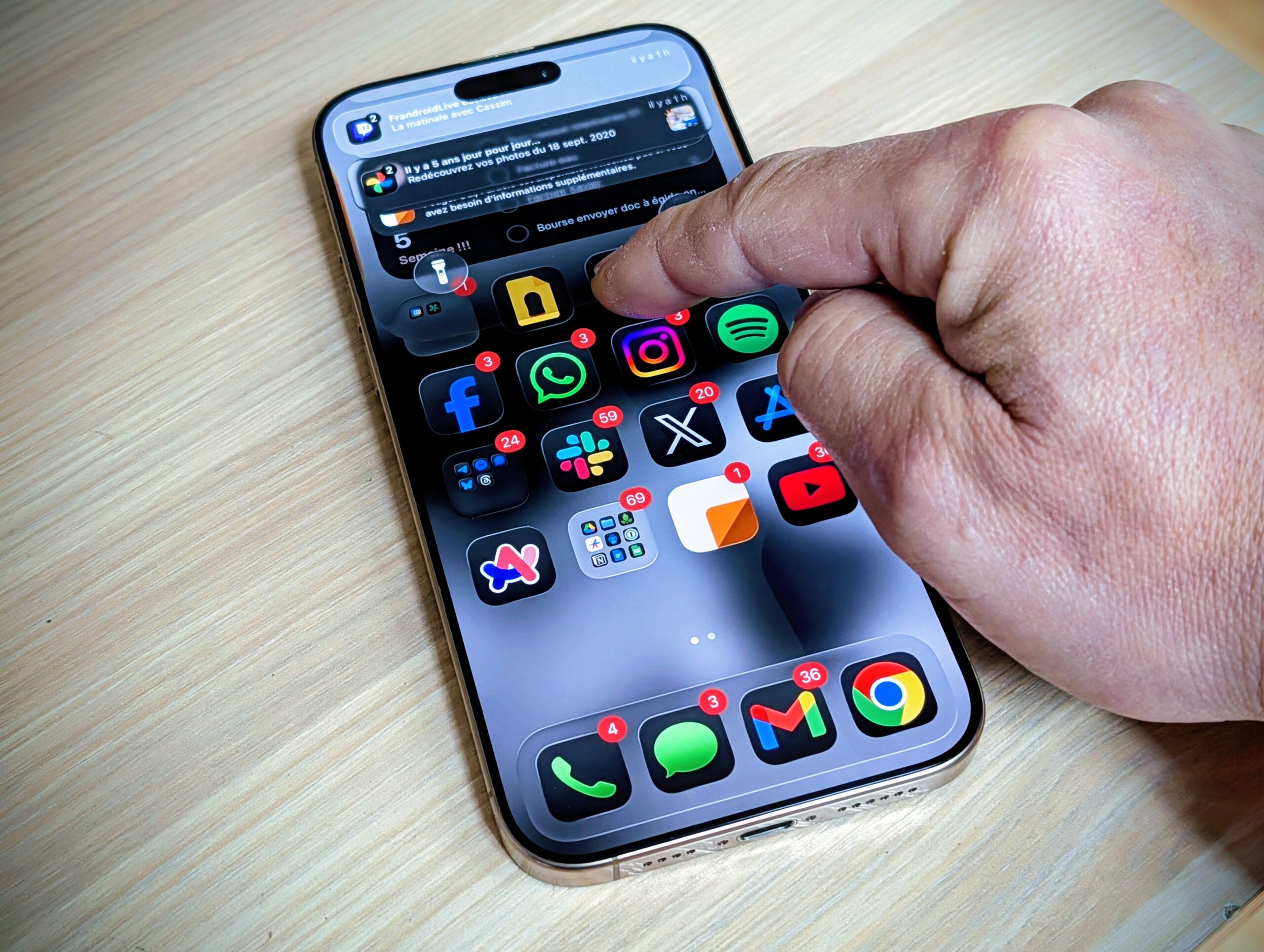 من موقع فون إسلام: تتفاعل اليد مع الهاتف الذكي الذي يعمل بنظام iOS 26، وتعرض العديد من أيقونات التطبيقات مع شارات الإشعارات على سطح خشبي، معززة بتقنية تقليل الشفافية لرؤية أوضح.