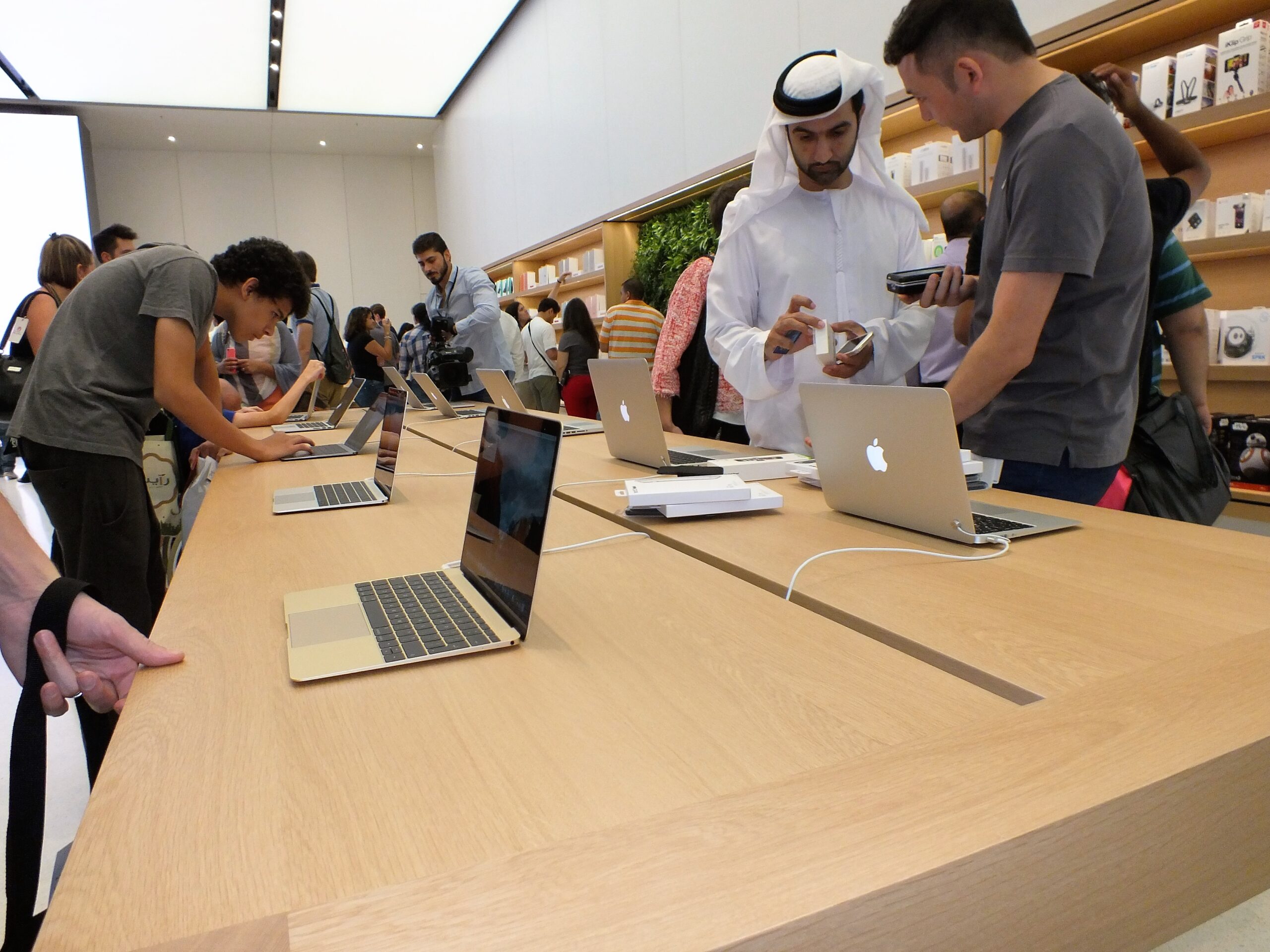 من موقع فون إسلام: العملاء يتصفحون ويختبرون أجهزة اللابتوب والإلكترونيات في متجر عصري مع طاولات خشبية ورف أن تعرض المنتجات في أجواء مستوحاة من أخبار التقنية لهذا على الهامش.