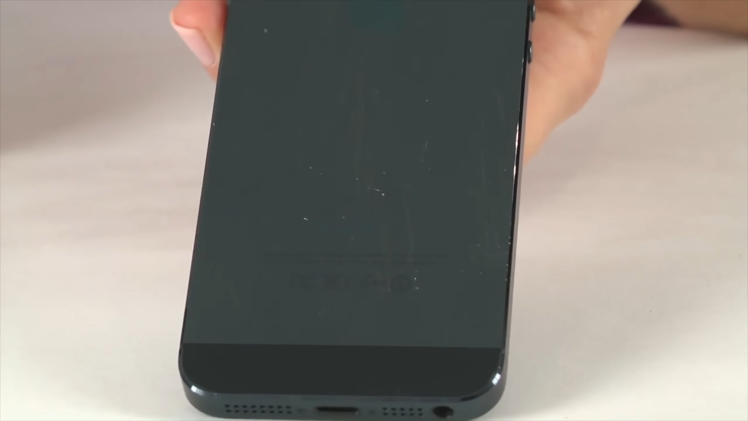 من موقع فون إسلام: يد تحمل هاتفًا ذكيًا أسود اللون بشاشة مخدوشة - مثال على أخطاء آبل - تظهر الحافة السفلية مع منفذ الشحن وشبكات مكبرات الصوت.