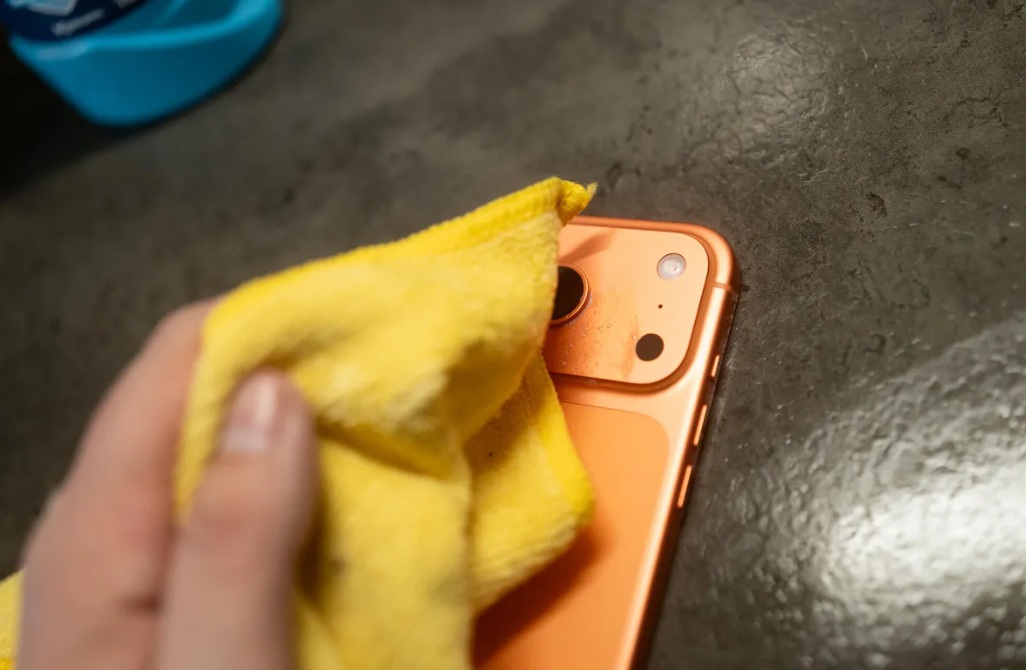 من موقع فون إسلام: يقوم شخص بتنظيف عدسة كاميرا أي- 17 برو البرتقالي بقطعة قماش صفراء على سطح مظلم.