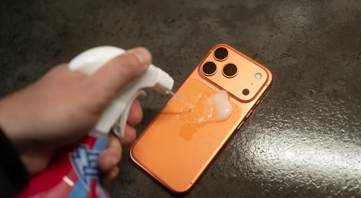 من موقع فون إسلام: تقوم اليد برش محلول التنظيف على ظهر أي- 17 برو البرتقالي موضوع على سطح عمل داكن.