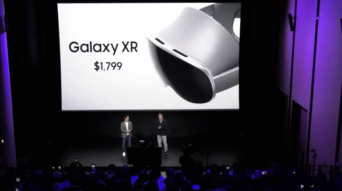 من موقع فون إسلام: يقف اثنان من مقدمي العروض على خشبة المسرح أمام شاشة تعرض "Galaxy XR بقيمة 1799 دولارًا أمريكيًا" وصورة لـ سامسونج نظارة جلاكسي XR، مستعرضين أحدث تقنيات الواقع الممتد.