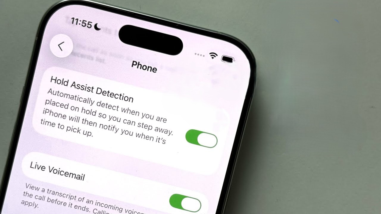 من موقع فون إسلام: تعرض شاشة الهاتف الذكي الذي يعمل بنظام iOS 26 إعدادات مساعد الانتظار (ميزة التعليق المساعد) والبريد الصوتي المباشر، وكلاهما تم تشغيلهما ضمن قائمة الهاتف. الوقت المعروض هو 11:55.