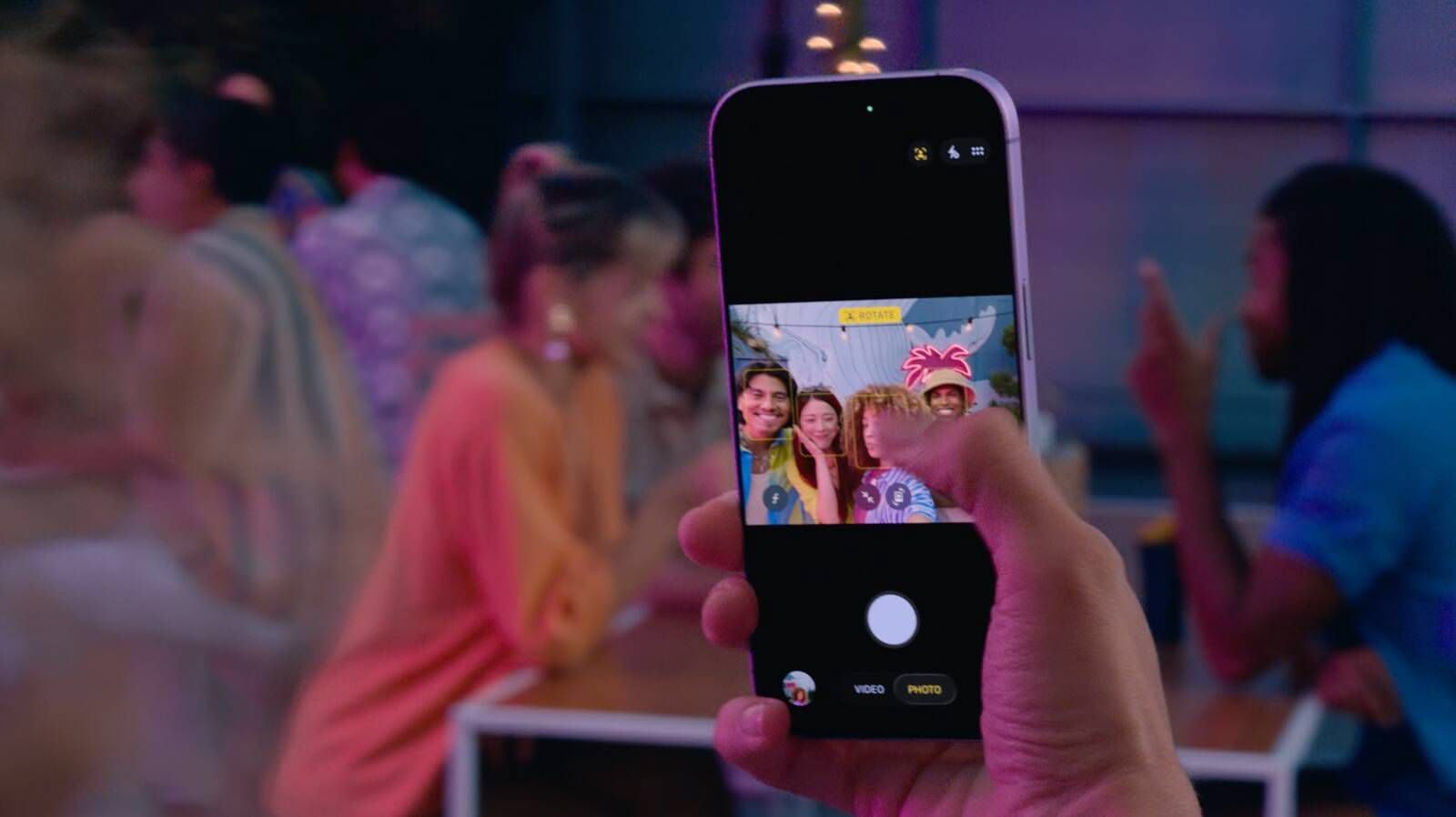 من موقع فون إسلام: شخص يستخدم كاميرا آي-فون 17 مع كاميرا سنتر ستيج لالتقاط صورة لثلاثة أصدقاء يبتسمون ويقفون في تجمع اجتماعي في الهواء الطلق خلال المساء.