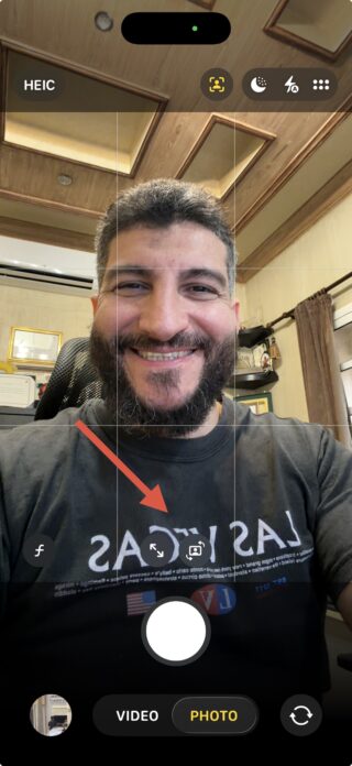 من موقع فون إسلام: رجل يبتسم في صورة سيلفي على شاشة تطبيق كاميرا الهاتف الذكي، مستفيداً من مركز الصورة. يشير سهم أحمر إلى أيقونة قلب الكاميرا. تتميز الغرفة بألواح خشبية ورفوف في الخلفية.