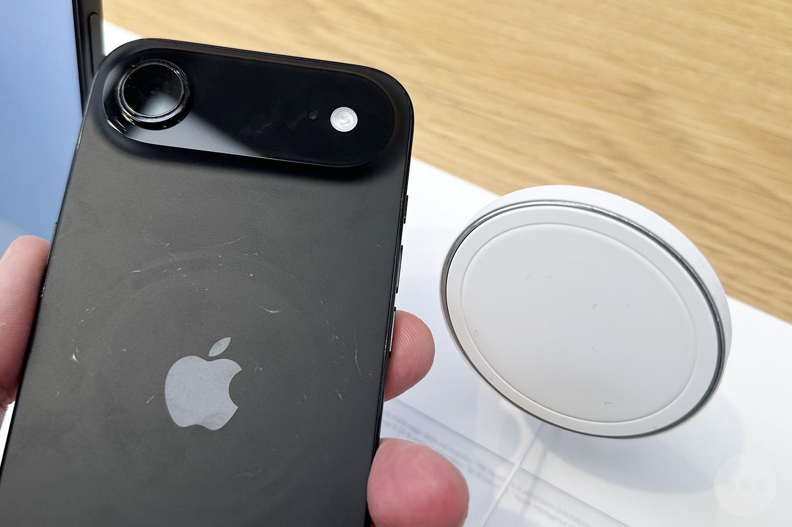 من موقع فون إسلام: شخص يحمل هاتف آيفون أسود اللون مع علامة دائرية على ظهره بجانب لوحة شحن لاسلكية بيضاء على سطح خشبي، في صورة أخبار التقنية على الهامش في سبتمبر.