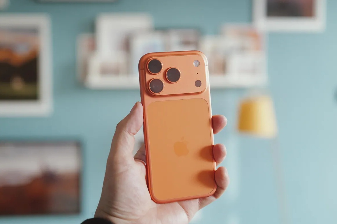 من موقع فون إسلام: يد تحمل برو برو 17 برو البرتقالي مع ثلاث كاميرات خلفية، على جدار أزرق غير واضح مزين بصور ذات أطر ومصباح أصفر في الخلفية.