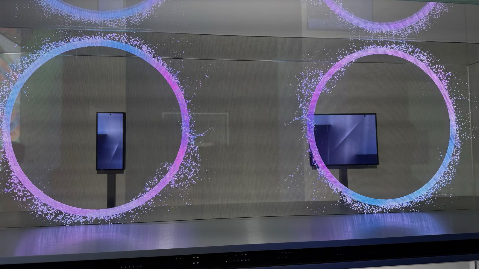 من موقع فون إسلام: يتم عرض هاتفين ذكيين في وضع مستقيم خلف الزجاج، كل منهما محاط بحلقة رقمية من الجسيمات الملونة، مع تسليط الضوء على ملخص أخبار التقنية للأسبوع 24 - 30 أكتوبر على منصة بسيطة.