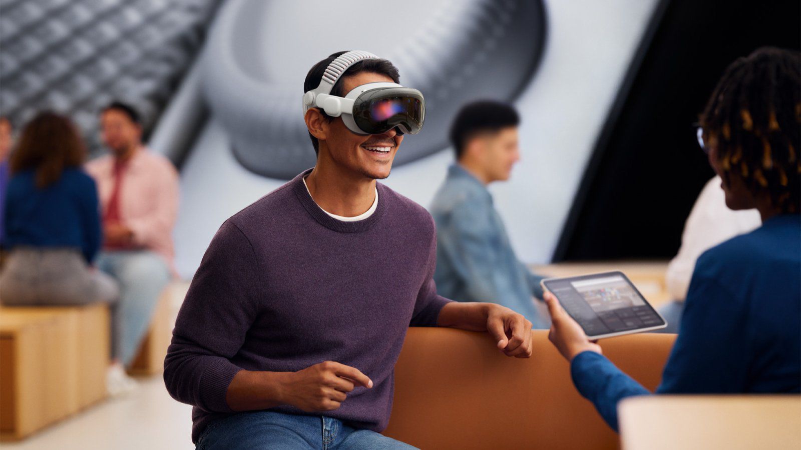 من موقع فون إسلام: رجل يرتدي سماعة واقع افتراضي يجلس ويبتسم أثناء تفاعله مع شخص آخر يحمل جهازًا لوحيًا في مكان عصري غير رسمي، ويناقش أخبار التقنية الحديثة وأحدث الابتكارات على الهامش في أكتوبر.