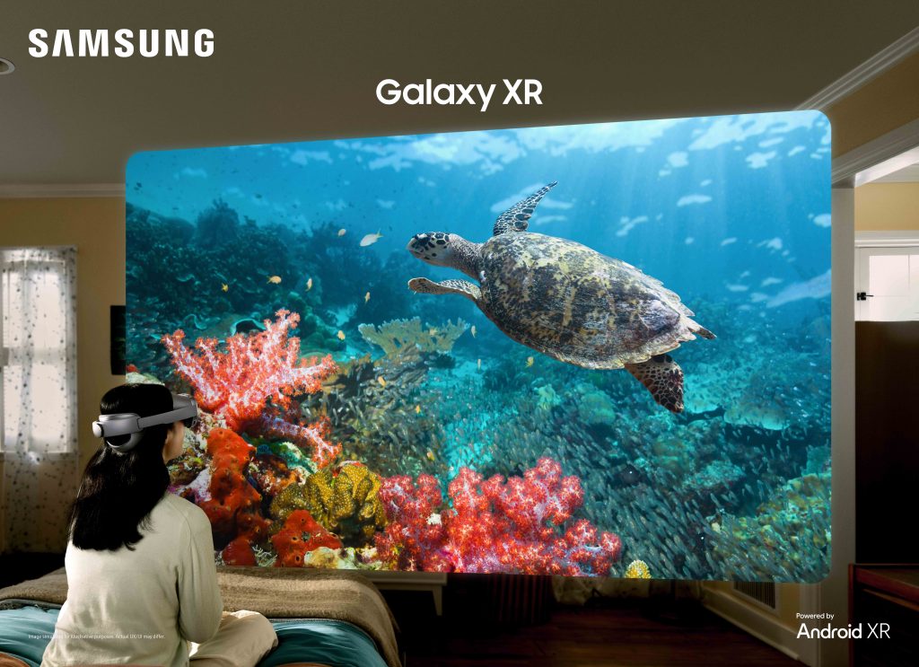 من موقع فون إسلام: شخص يرتدي نظارة سامسونج سامسونج XR يجلس على سرير ويشاهد مشهداً افتراضياً تحت الماء مع سلحفاة بحرية ومرجان معروض في غرفته. وتظهر شعارات Samsung وGalaxy XR، مما يجعلها مثالية لمقارنة تجربة الواقع الافتراضي.