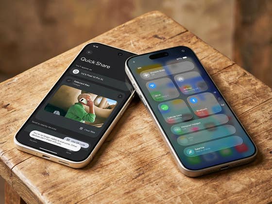 من موقع فون إسلام: يستند هاتفان ذكيان على سطح خشبي؛ يعرض أحدهما شاشة "المشاركة السريعة"، بينما يعرض الآخر لوحة الاتصال والإعدادات - تسليط الضوء على نقل الملفات بين آي أون وأندرويد.