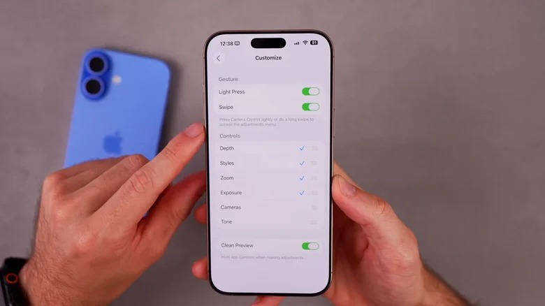 من موقع فون إسلام: شخص يحمل جهاز آي فون يعرض إعدادات التخصيص، مع إبراز ميزة كاميرا جديدة بينما يستقر جهاز آي فون أزرق على طاولة في الخلفية.