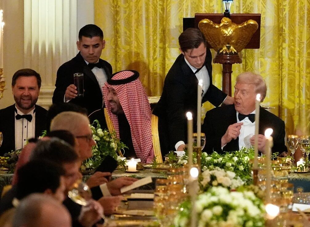 من موقع فون إسلام: مشهد عشاء رسمي مع العديد من الضيوف الجالسين على مائدة طويلة، وخدم يحضرون الطعام، وإعدادات مائدة متقنة, قطعة مركزية مزخرفة على شكل نسر ذهبي في الخلفية، ومناقشة هادئة حول أخبار أخبار التقنية و ملخص الأسبوع.