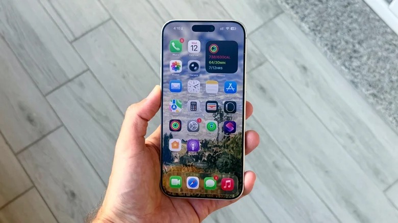 Von PhoneIslam: Eine Hand, die ein Smartphone hält, zeigt einen Startbildschirm mit verschiedenen App-Symbolen und einer beeindruckenden Landschaftskulisse, die die Funktionen des iOS-Betriebssystems hervorhebt. Im Hintergrund sind ein gefliester Boden und eine strukturierte Oberfläche zu sehen.