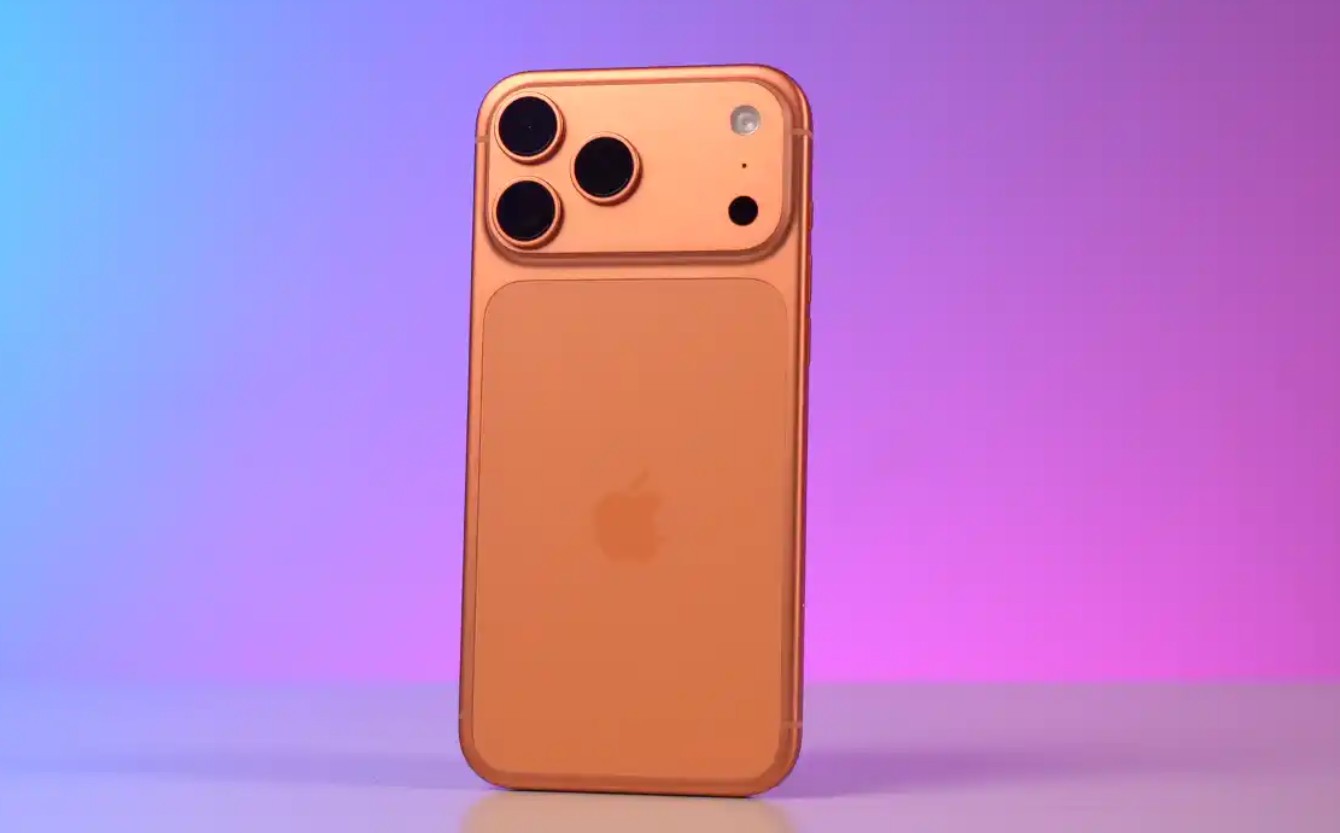 من موقع فون إسلام: آي-فون 17 برو بلون ذهبي مع ثلاث كاميرات خلفية تقف في وضع مستقيم على خلفية متدرجة باللونين الأرجواني والوردي، في انتظار الشحن وسط ضوضاء ضوئية.