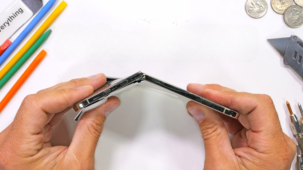 من موقع فون إسلام: شخص يثني هاتفًا ذكيًا من طراز آي-فون إير إلى النصف، مسببًا تلفًا واضحًا، مع وجود أدوات وأقلام رصاص ملونة وعملات معدنية مبعثرة على سطح أبيض بالقرب منه.