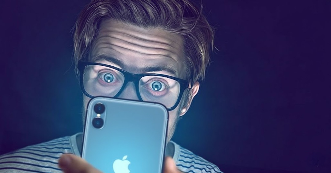 من موقع فون إسلام: شخص يرتدي نظارة يحدق بعيون واسعة في هاتف ذكي متوهج في مكان خافت الإضاءة، مما يبرز تأثيرات اجتهاد العين.