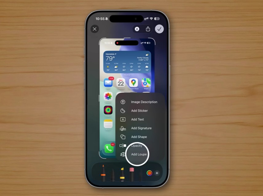 من موقع فون إسلام: يعرض الهاتف الذكي الذي يعمل بنظام iOS 26 قائمة تحرير لقطة شاشة مع خيارات مثل وصف الصورة، وإضافة ملصق، وإضافة نص، وإضافة توقيع مقابل ميزات خفية على سطح خشبي يبرز ميزات خفية على الآي-فون.
