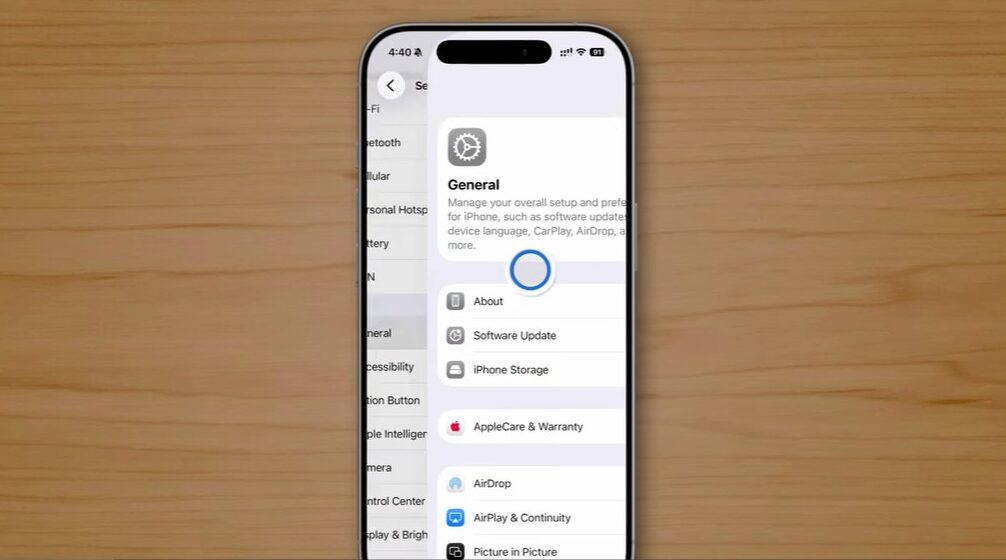 من موقع فون إسلام: هاتف ذكي يعرض قائمة الإعدادات "العامة" على سطح خشبي، مع خيارات مثل حول، وتحديث البرنامج، وتخزين الهاتف، وهي خيارات مرئية على الشاشة - مثالية لاستكشاف ميزات خفية في نظام التشغيل iOS 26 على الآي فون.