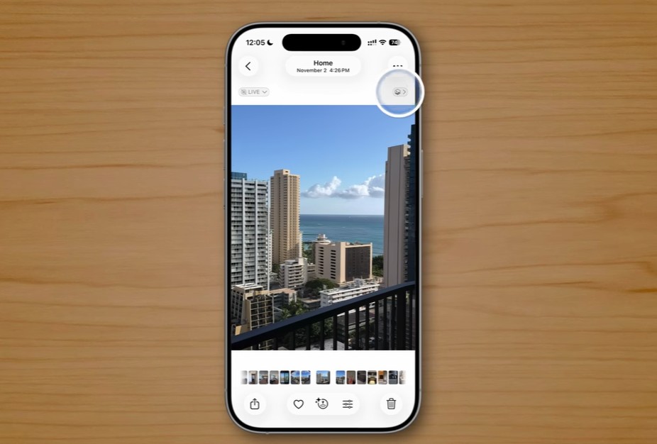 من موقع فون إسلام: هاتف ذكي، مثل الآي فون، يعرض صورة لمباني المدينة والمحيط على طاولة خشبية، مع تمييز أيقونة في الشاشة لعرض ميزات خفية من iOS 26.