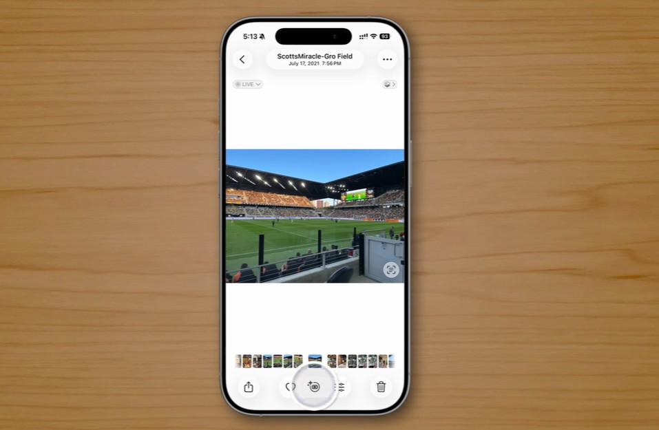 من موقع فون إسلام: يعرض نظام التشغيل iOS 26 الآي-فون صورة لملعب كرة قدم على شاشته، مع وضع الهاتف على سطح خشبي خفيف.