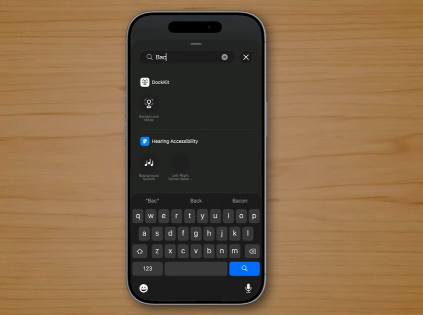 من موقع فون إسلام: يعرض الهاتف الذكي على سطح خشبي شريط بحث مع كتابة كلمة "باك"، وتظهر نتائج البحث ولوحة مفاتيح في الأسفل، مع تسليط الضوء على ميزات خاتفية جديدة في الآي-فون مع iOS 26.