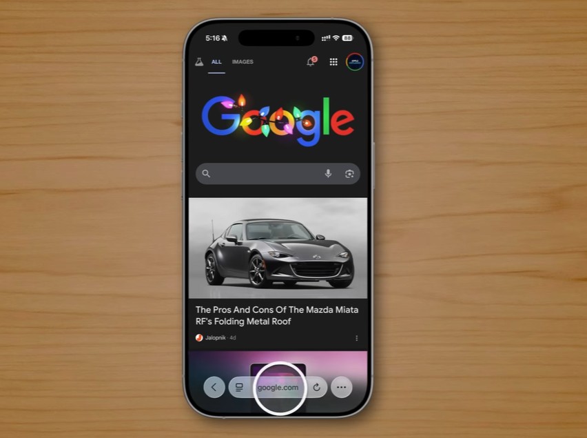 من موقع فون إسلام: هاتف ذكي يعمل بنظام iOS 26 يعرض الصفحة الرئيسية لجوجل مع نتيجة بحث عن سيارة مازدا مياتا، موضوعة على سطح خشبي، ويكشف عن ميزات خفية في الآي-فون.