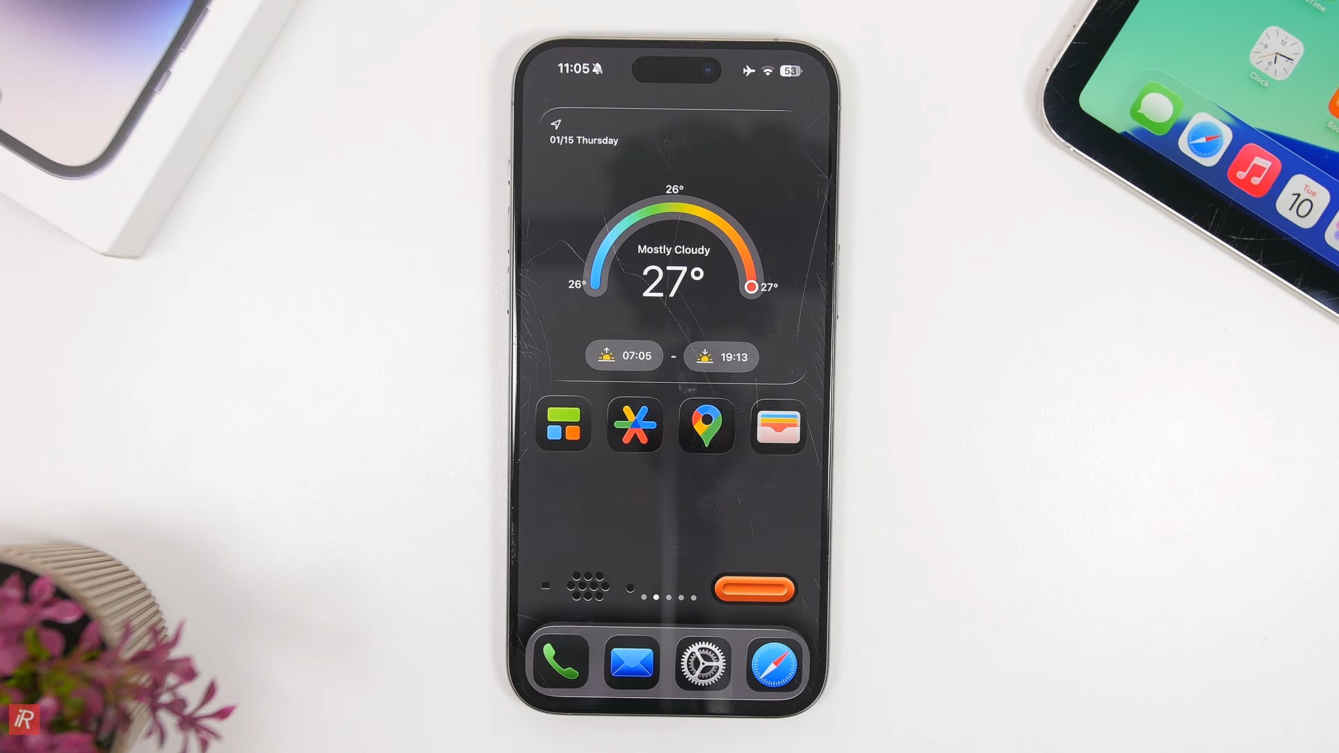 من موقع فون إسلام: جهاز آي-فون 17 يعرض أداة طقس والعديد من أيقونات التطبيقات على شاشته على سطح أبيض بجانب حافة جهاز لوحي ونبتة صغيرة في أصيص صغير، يوضح بعض حيل نظام iOS 26 المفيدة.