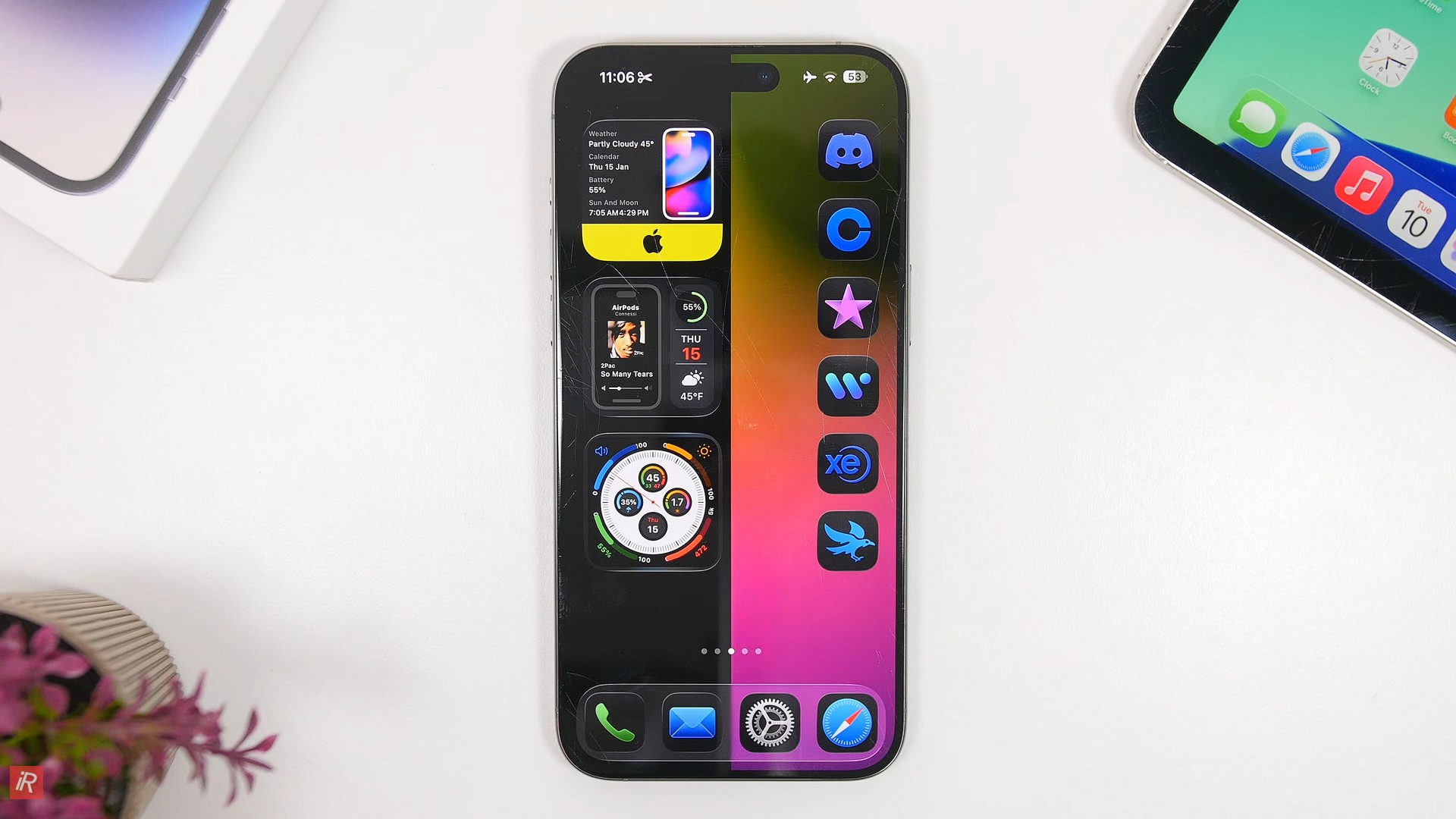 من موقع فون إسلام: يعرض هاتف ذكي يعمل بنظام iOS 26 شاشة رئيسية مخصصة مع العديد من أيقونات التطبيقات والأدوات المصغّرة الموضوعة على سطح أبيض بجانب جهاز لوحي ونبتة في أصيص.