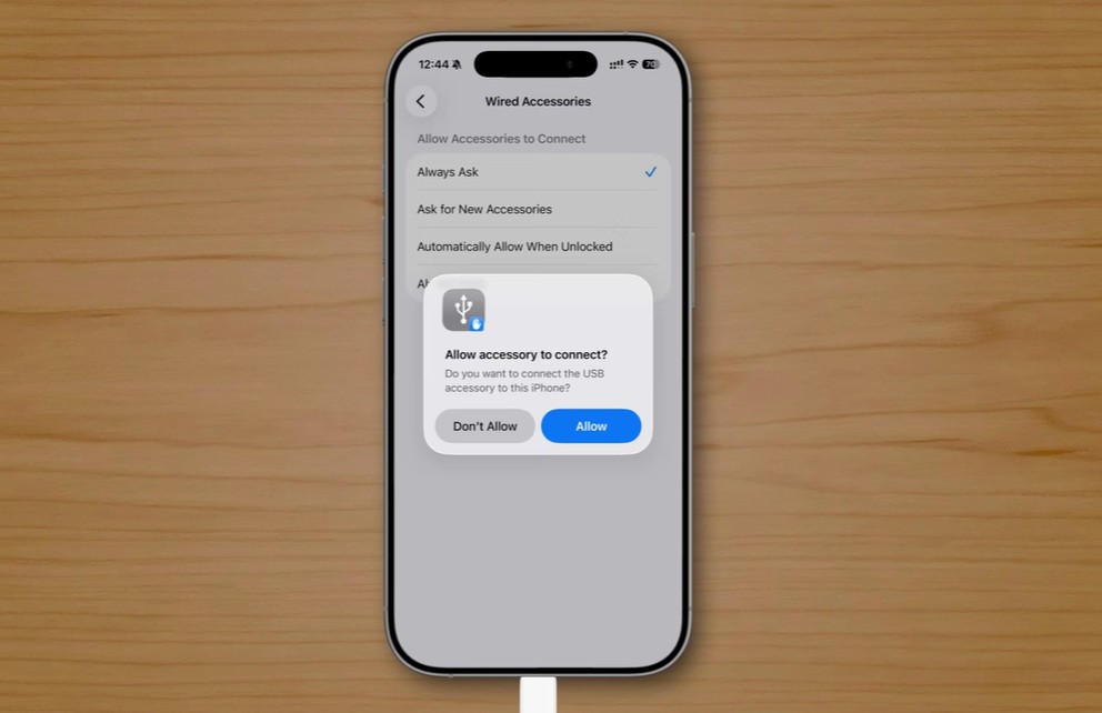 من موقع فون إسلام: تعرض نافذة منبثقة تطلب السماح بتوصيل ملحق USB، مع خيارات "عدم السماح" أو "السماح"، على خلفية طاولة خشبية - تعرض ميزات خفية تم تقديمها في نظام التشغيل iOS 26.