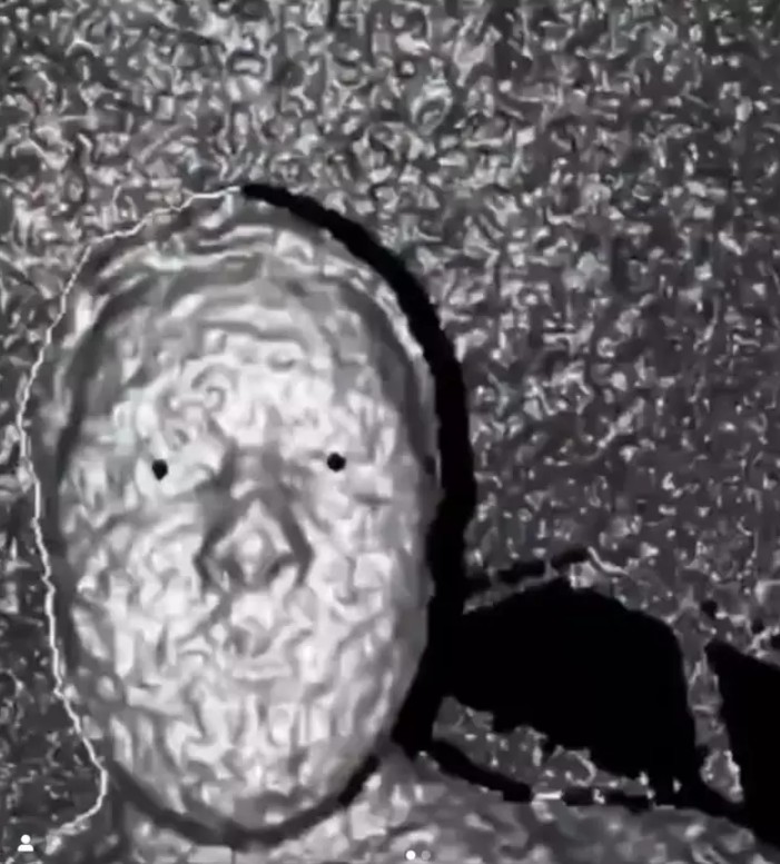 من موقع فون إسلام: يستحضر الوجه الرمادي المزخرف باللون الرمادي مع نقاط سوداء للعيون تأثيرًا تجريديًا مشوهًا - يذكرنا بـ كاميرا الآيكوني-فون وتخطيط الوجه على خلفية مزخرفة مماثلة.