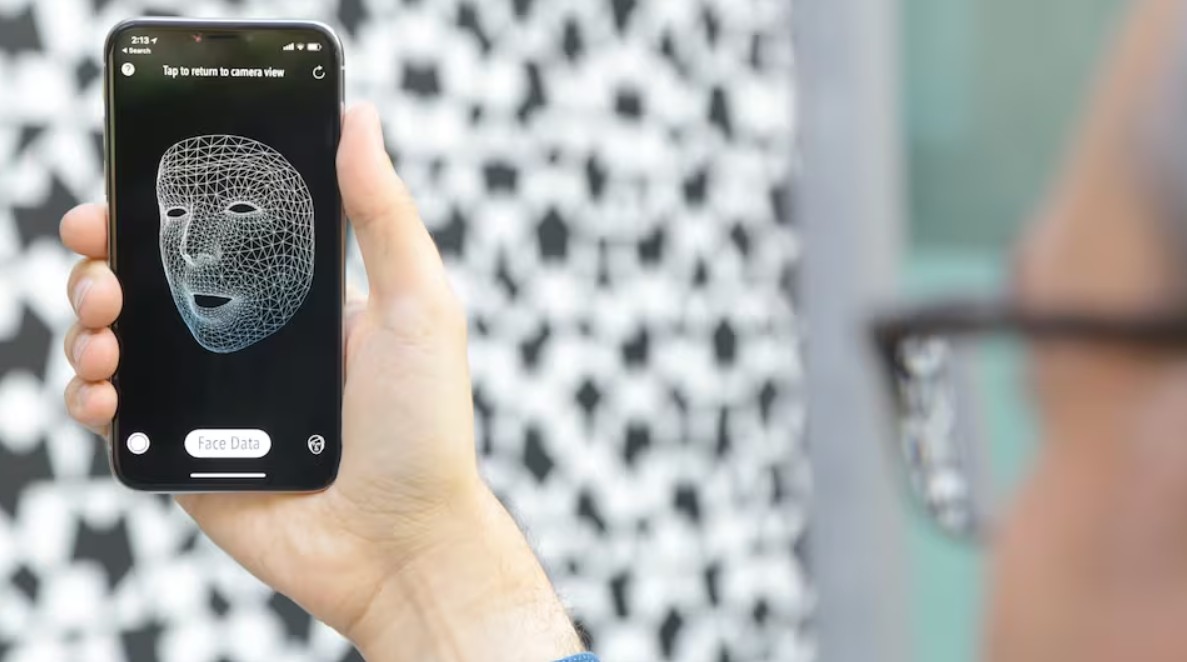 من موقع فون إسلام: شخص يحمل هاتفًا ذكيًا يعرض وجهًا شبكيًا رقميًا للتعرف على الوجه باستخدام Face ID، مع خلفية منقوشة غير واضحة.