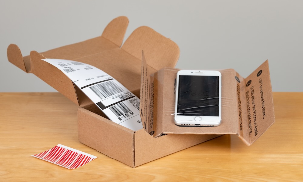 من موقع فون إسلام: هاتف ذكي أبيض بشاشة متصدعة، على الأرجح بسبب خطأ فادح, يوضع داخل صندوق شحن مفتوح مع ملصقات ظاهرة على طاولة خشبية - ربما في انتظار برنامج إعادة التعبئة والتغليف - بعد هذه العملية.