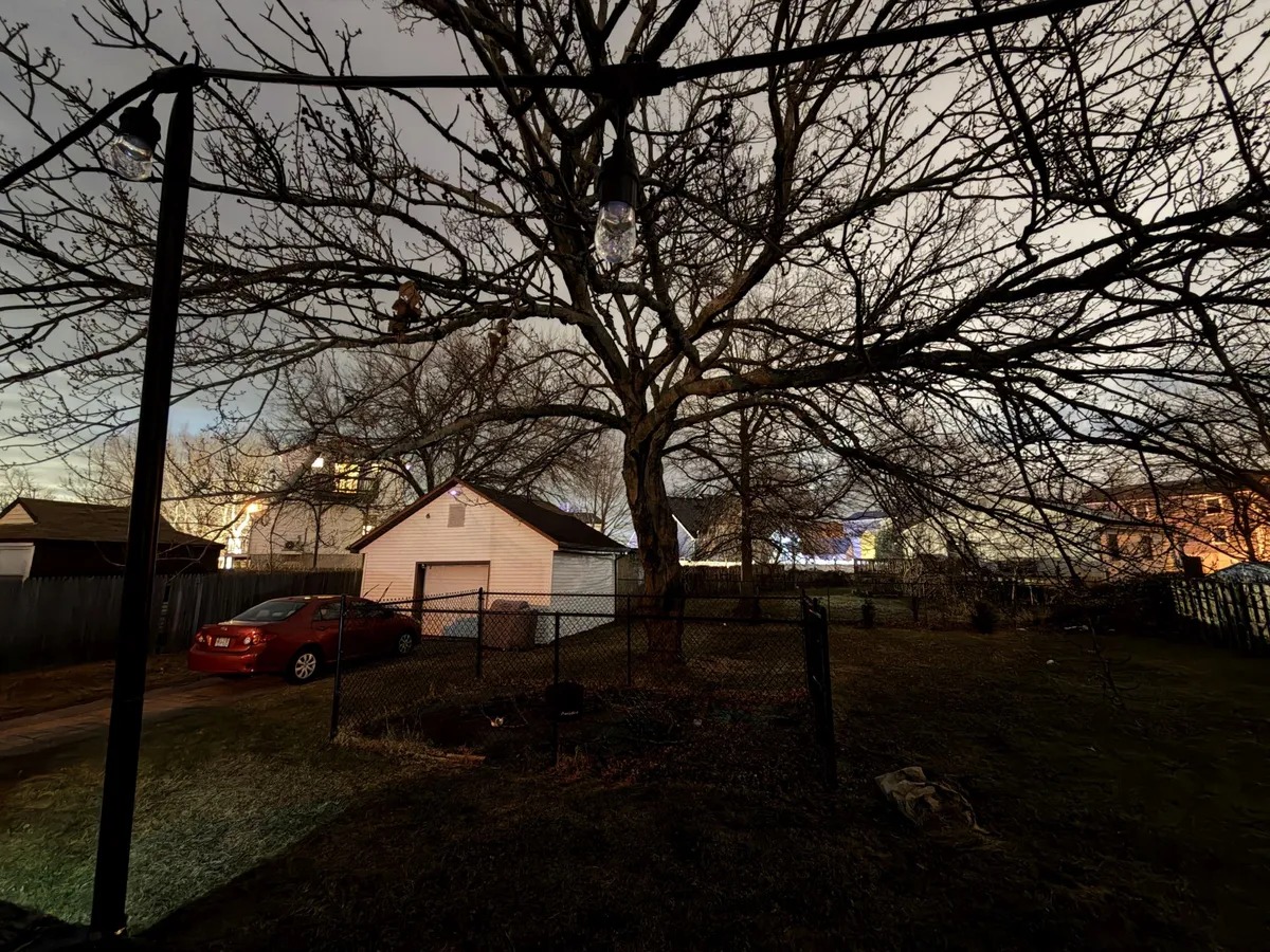 من موقع فون إسلام: يلتقط "الوضع الليلي ماكس" مشهداً ليلياً لفناء خلفي به شجرة كبيرة بلا أوراق وسيارة حمراء ومرآب أبيض وسياج تحت سماء ملبدة بالغيوم مع منازل تتوهج بهدوء في الخلفية.