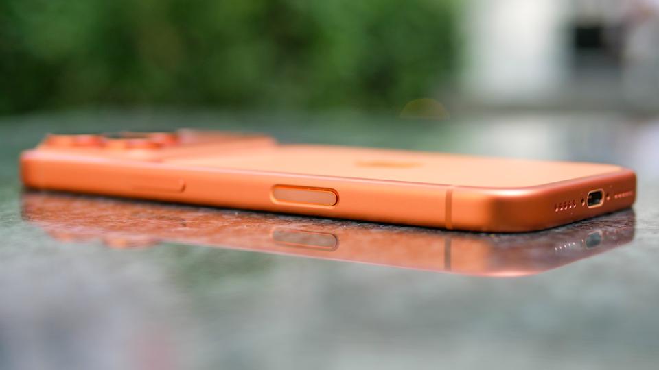 من موقع فون إسلام: هاتف ذكي برتقالي اللون مسطح على سطح عاكس، يظهر الزر الجانبي ومنفذ الشحن - هاتف ذكي برتقالي اللون مسطح على سطح عاكس - مع خلفية خضراء غير واضحة.