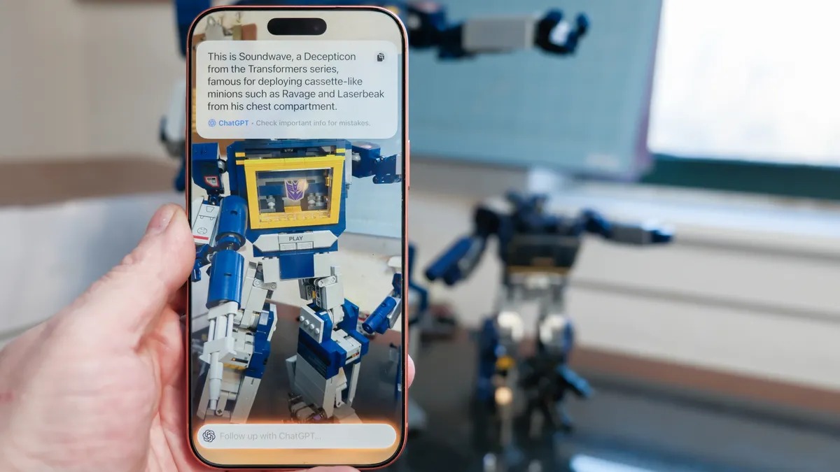 من موقع فون إسلام: يد تحمل هاتفًا ذكيًا يعرض صورة ووصفًا للعبة ترانسفورمرز ساوندويف ترانسفورمرز، مع تسليط الضوء على مزايا التحكم بالكاميرا في الآي آفون؛ ويظهر في الخلفية تمثال لعبة مطابق على طاولة.