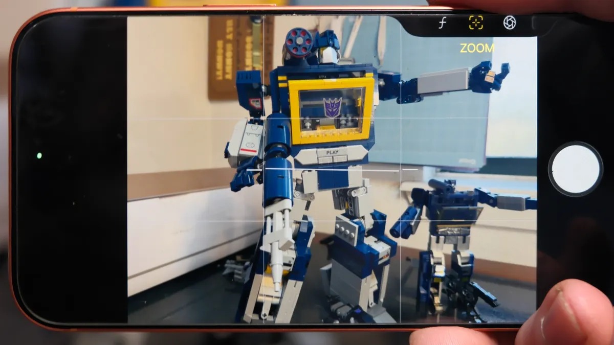 من موقع فون إسلام: تعرض شاشة الهاتف الذكي صورة يتم التقاطها لشخصيتين من شخصيات ليغو ترانسفورمرز موضوعتين على طاولة، وتبرز مزايا التحكم بالكاميرا على آي آيفون مع وجود شخصية في المقدمة وشخصية أخرى أصغر على اليمين.